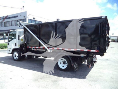 Isuzu NRR roll-off truck in a dealership lot with black dump body and white cab. Visible text includes no legible labeling on the body in this view.