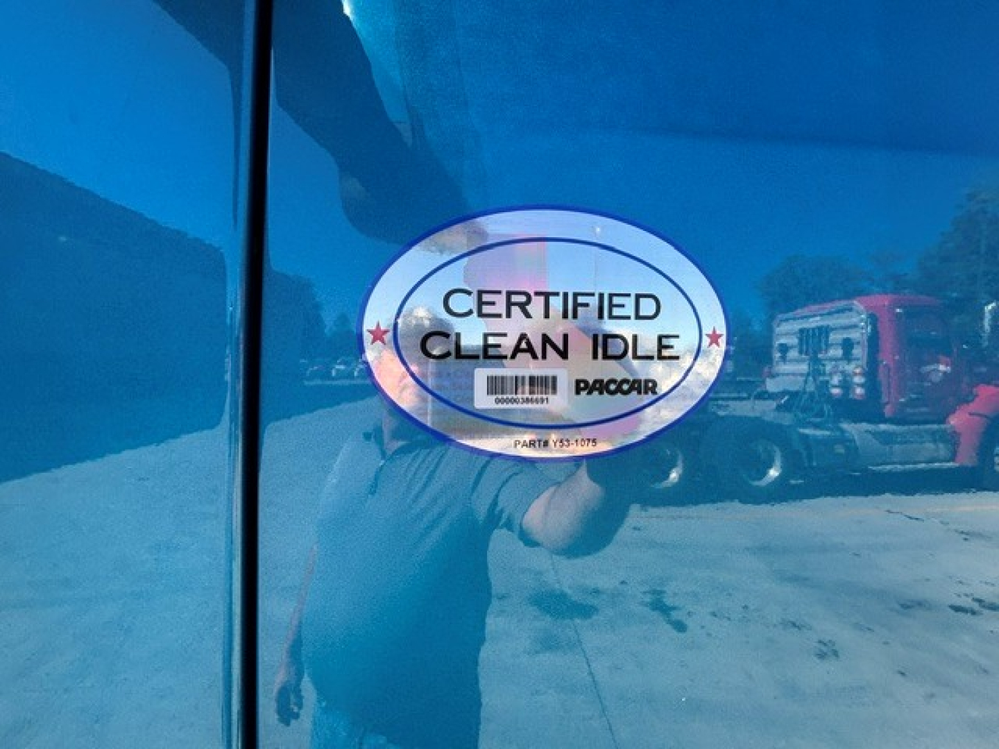 Sticker on blue Peterbilt door showing CERTIFIED CLEAN IDLE, with PACCAR logo and barcode; reflection of person taking photo.