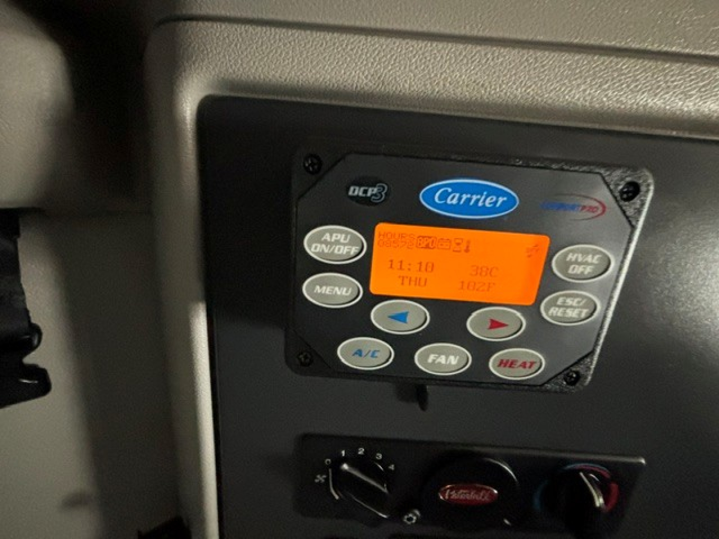 Carrier/Bluedog vehicle climate control panel with digital display and A/C controls inside a Peterbilt cab. Visible text includes Carrier logo, APU ON/OFF, MENU, A/C, DEF, HEAT, and numeric readout.