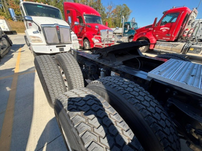 Peterbilt 579 chassis view showing dual rear axles and tires, with nearby white and red trucks in a dealership lot.