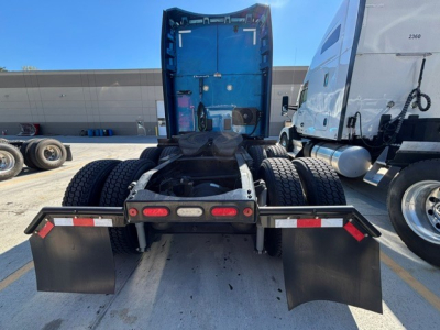 Peterbilt truck rear view showing fifth wheel, two rear axles, mud flaps, and tail lights with blue cab in background.