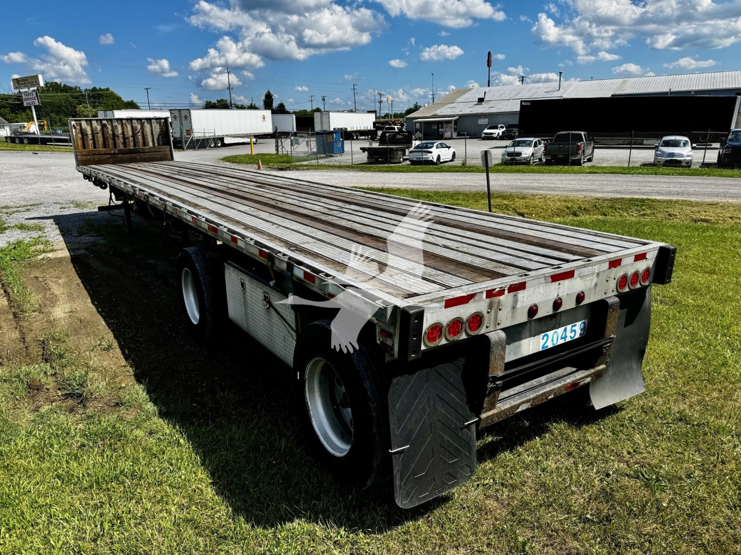 Flatbed trailer with open deck and dual rear axles, back end showing taillights and mud flaps; aluminum deck with wood side stakes in a grassy lot.
