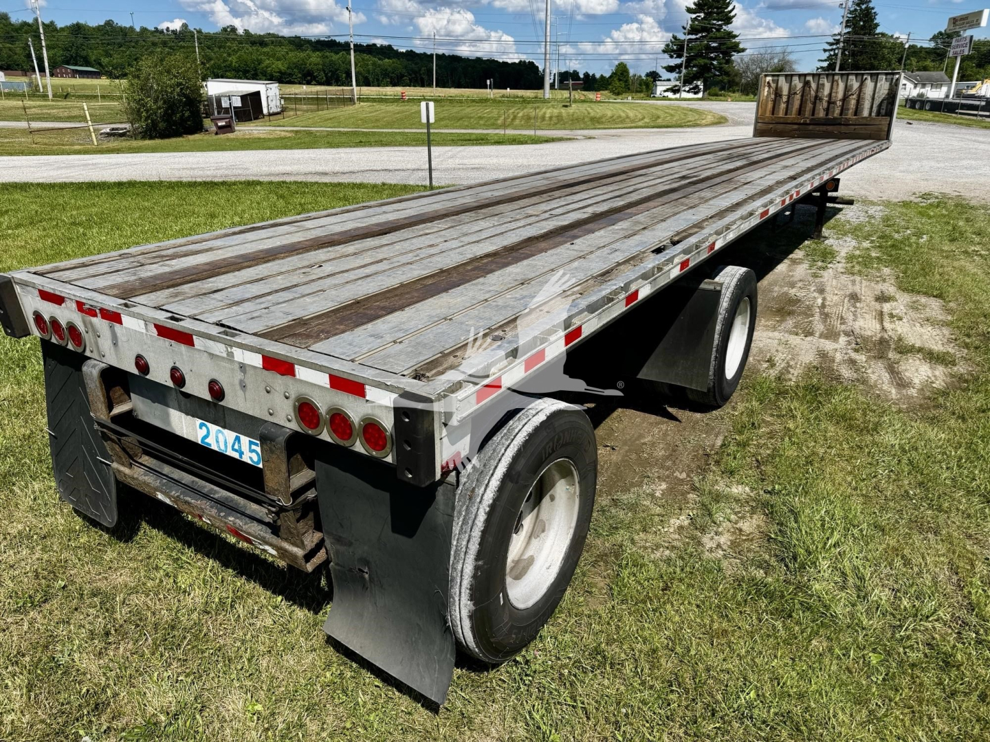 Flatbed trailer showing weathered steel deck and dual rear axles with white wheel rims. Tail has 2045 plate, lights and reflectors along sides.