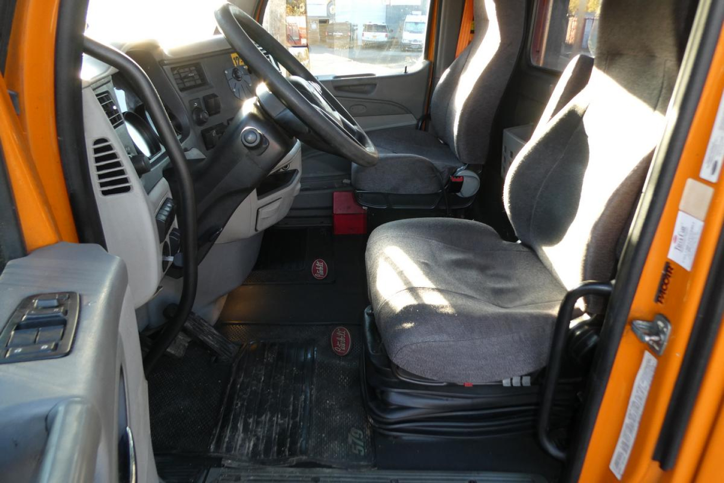 Truck cab interior with grey driver's seat and steering wheel visible; orange exterior frame; dashboard controls and pedals visible.