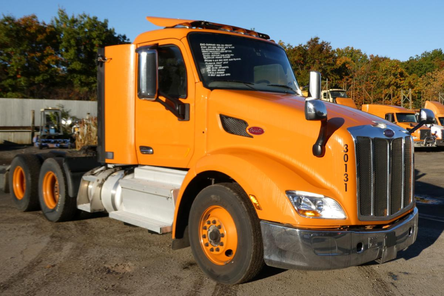 Peterbilt 579 daycab truck in orange, front-left view, showing cab, mirrors, and front grille; truck number 30131 on door; windshield has white text; heavy-duty tires; background with other trucks.