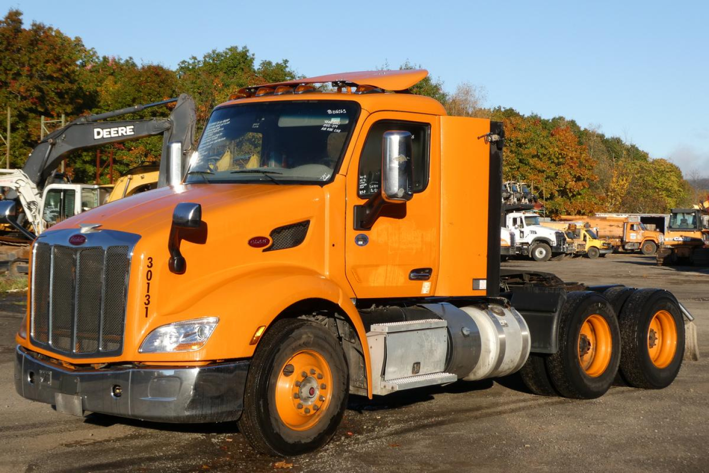 Orange Peterbilt 579 day cab semi-truck with front chrome grille, large mirrors, and dual rear axles; visible equipment and trucks in background.