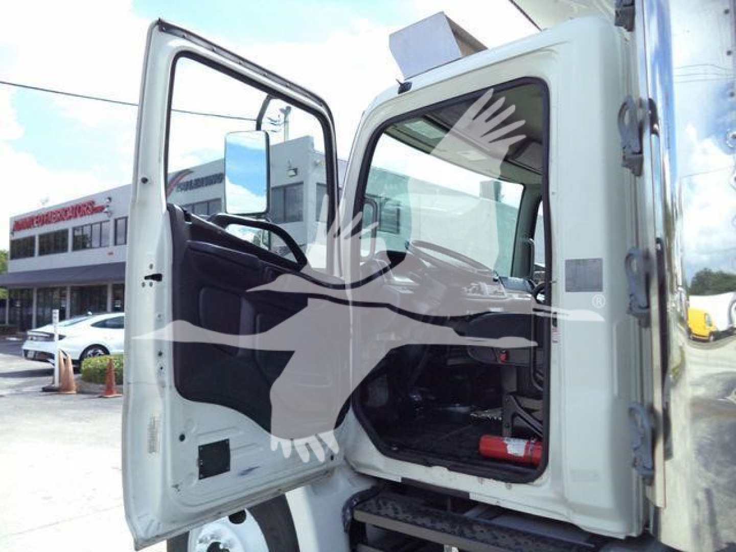 Truck cab interior visible through open door; steering wheel and driver seat; exterior white refrigerated truck cab splash and window; part of dealership lot in background.