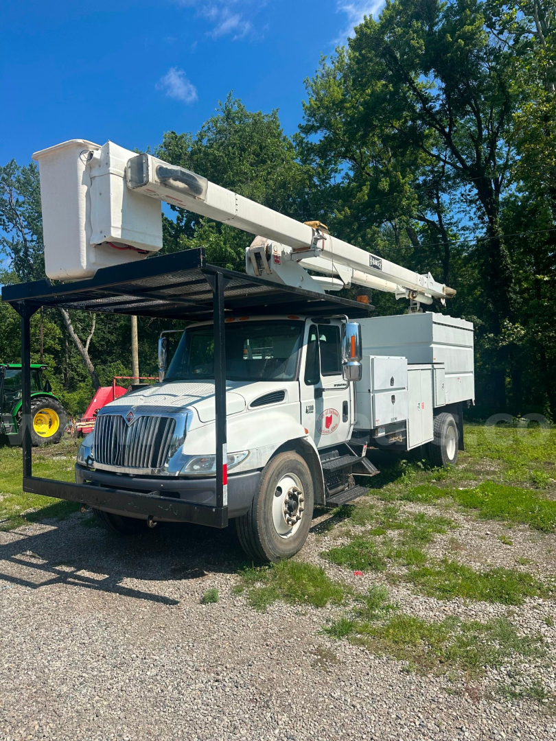 Bucket truck with extended boom mounted on a white International 4300 cab; utility compartments along the chassis; no visible cab damage; green trees in background.