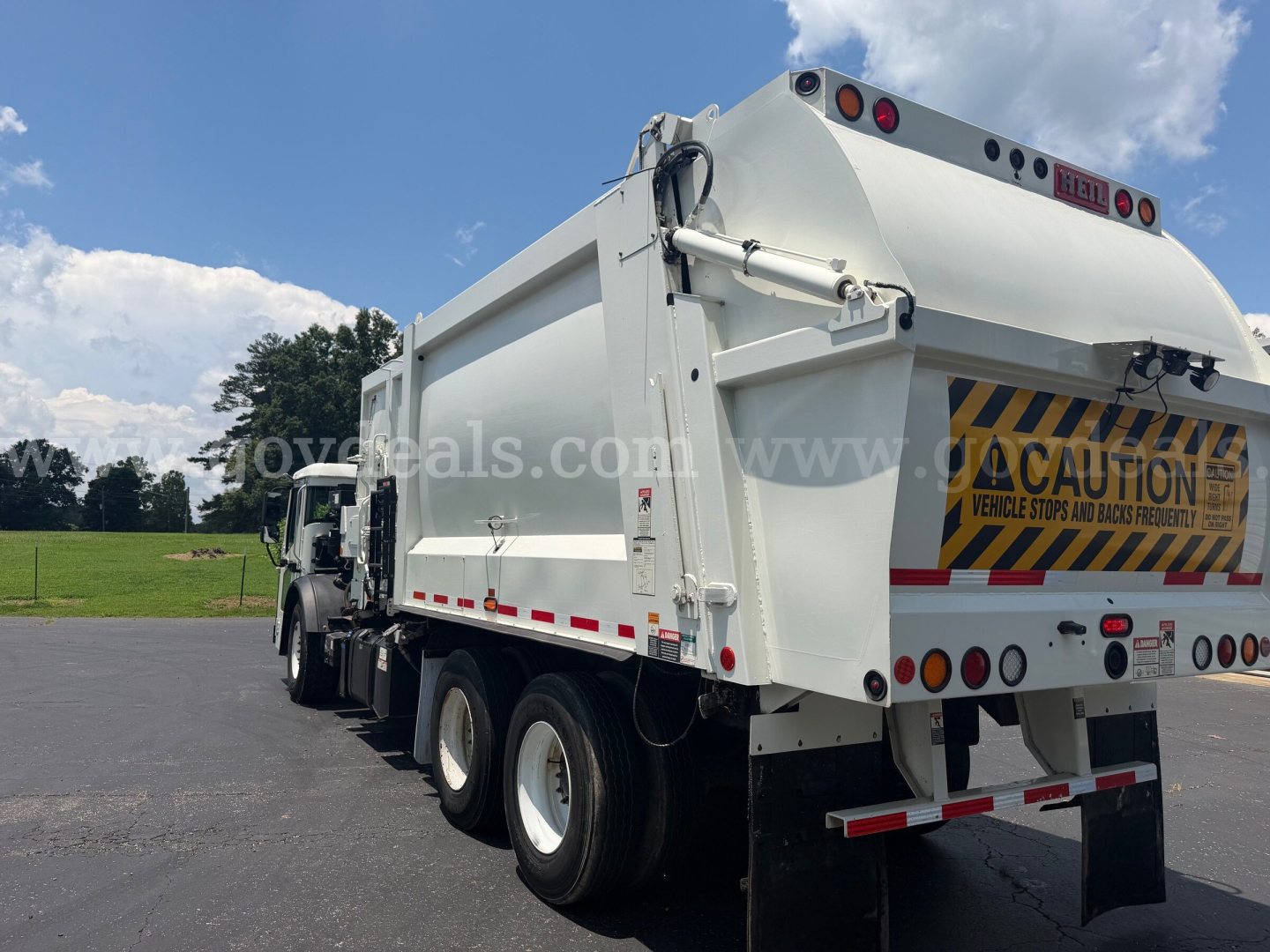 Rear/right side view of a white Mack LR dump truck in a parking lot, with warning decal and reflective stripes on the tail; visible are dual rear axles, dump bed, and CAUTION sign on tailgate.
