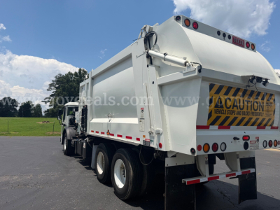 Rear/right side view of a white Mack LR dump truck in a parking lot, with warning decal and reflective stripes on the tail; visible are dual rear axles, dump bed, and CAUTION sign on tailgate.
