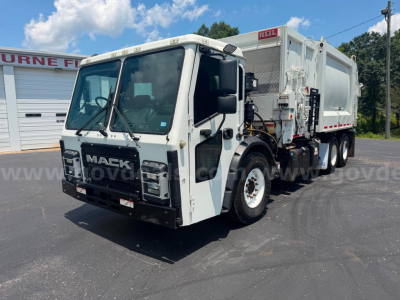 Garbage truck front-left view showing Mack branding on grille and cab, heavy-duty axles, side hopper, warning labels, and rear mixer with hydraulic lines.