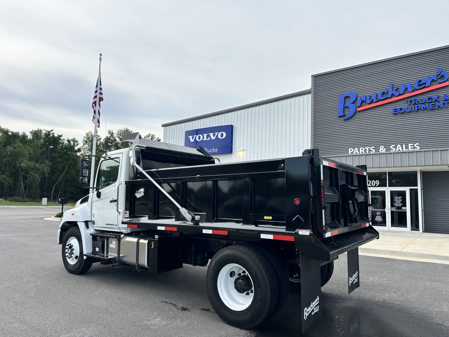 Truck bed area of a 2025 Hino L7 cab and chassis showing dump bed and rearview with wheel, front cab, and dealership building in background.