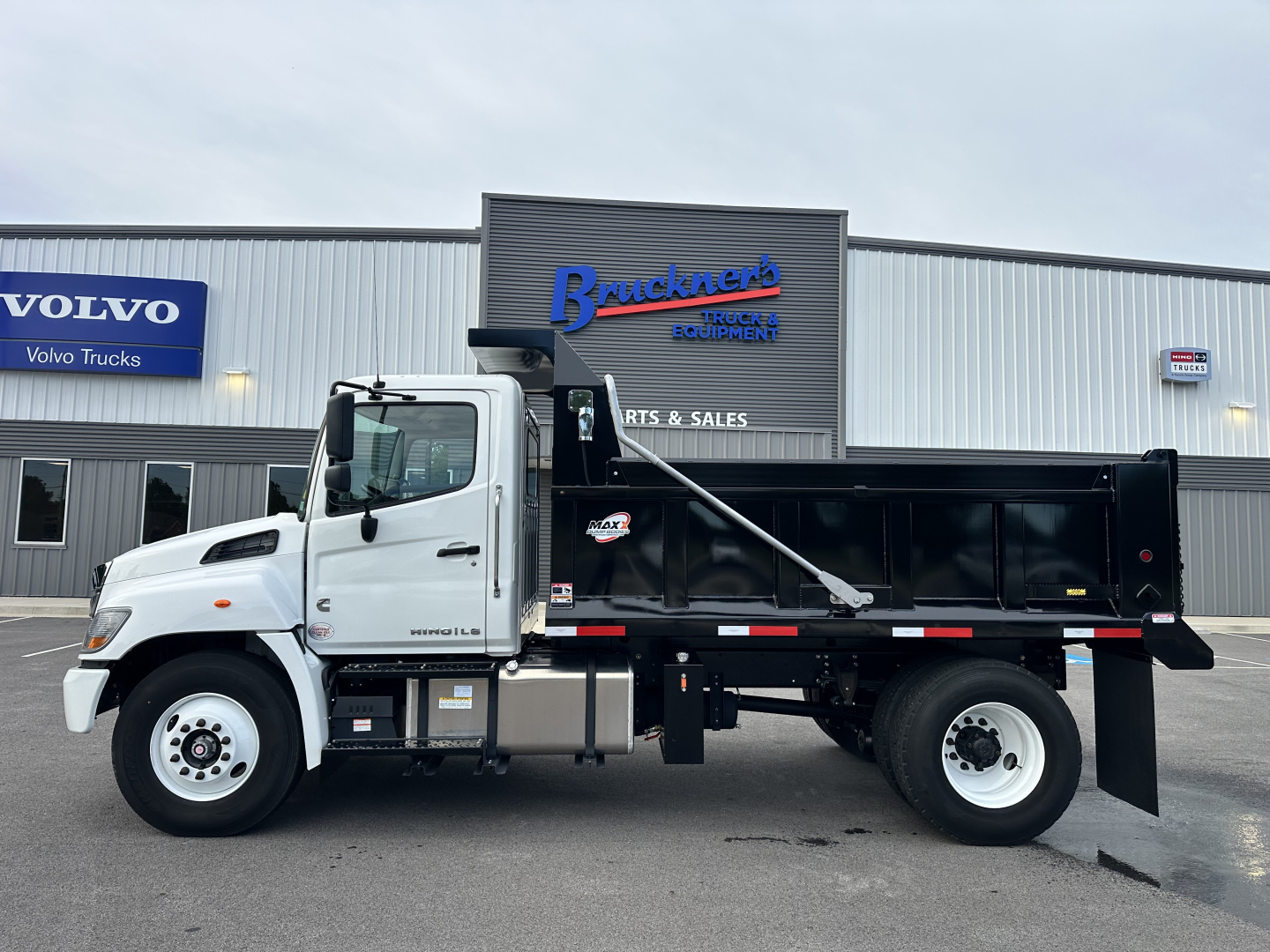 Front-left side view of a white hydraulic dump truck with a black dump bed, parked in front of a dealership. Visible text includes 'Bruckner's Equipment' on the building signage, 'VOLVO Trucks' on a blue sign, and 'DUMP' logo on the side of