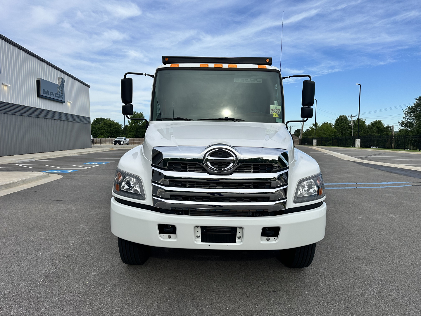 Front view of a 2025 Hino L7 cab and chassis truck showing chrome grille, large headlights, and Hino emblem; clear windshield, side mirrors, and amber cab roof lights.