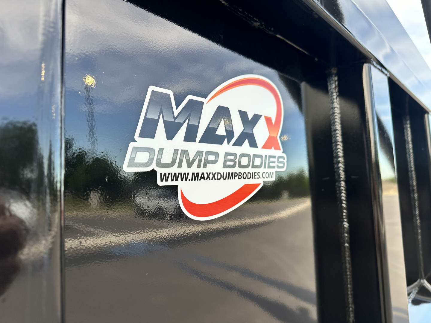Sticker label on a Max Dump Bodies equipment showing MAXX DUMP BODIES logo with web address on a black metal surface.