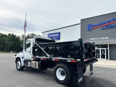 Truck bed area of a 2025 Hino L7 cab and chassis showing dump bed and rearview with wheel, front cab, and dealership building in background.