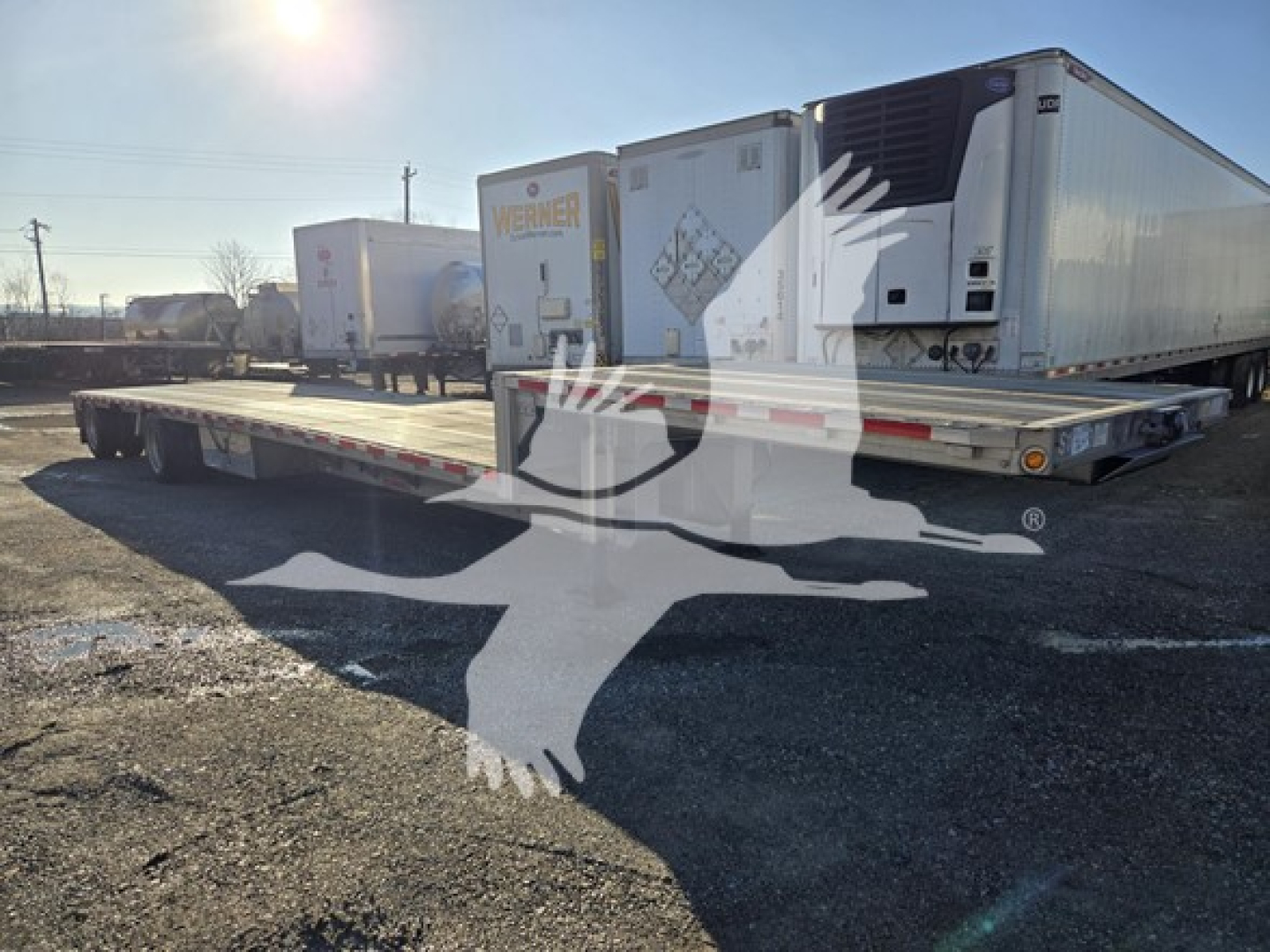 Drop deck trailer showing long flatbed with dual tandem axles and a white refrigerated trailer in background.