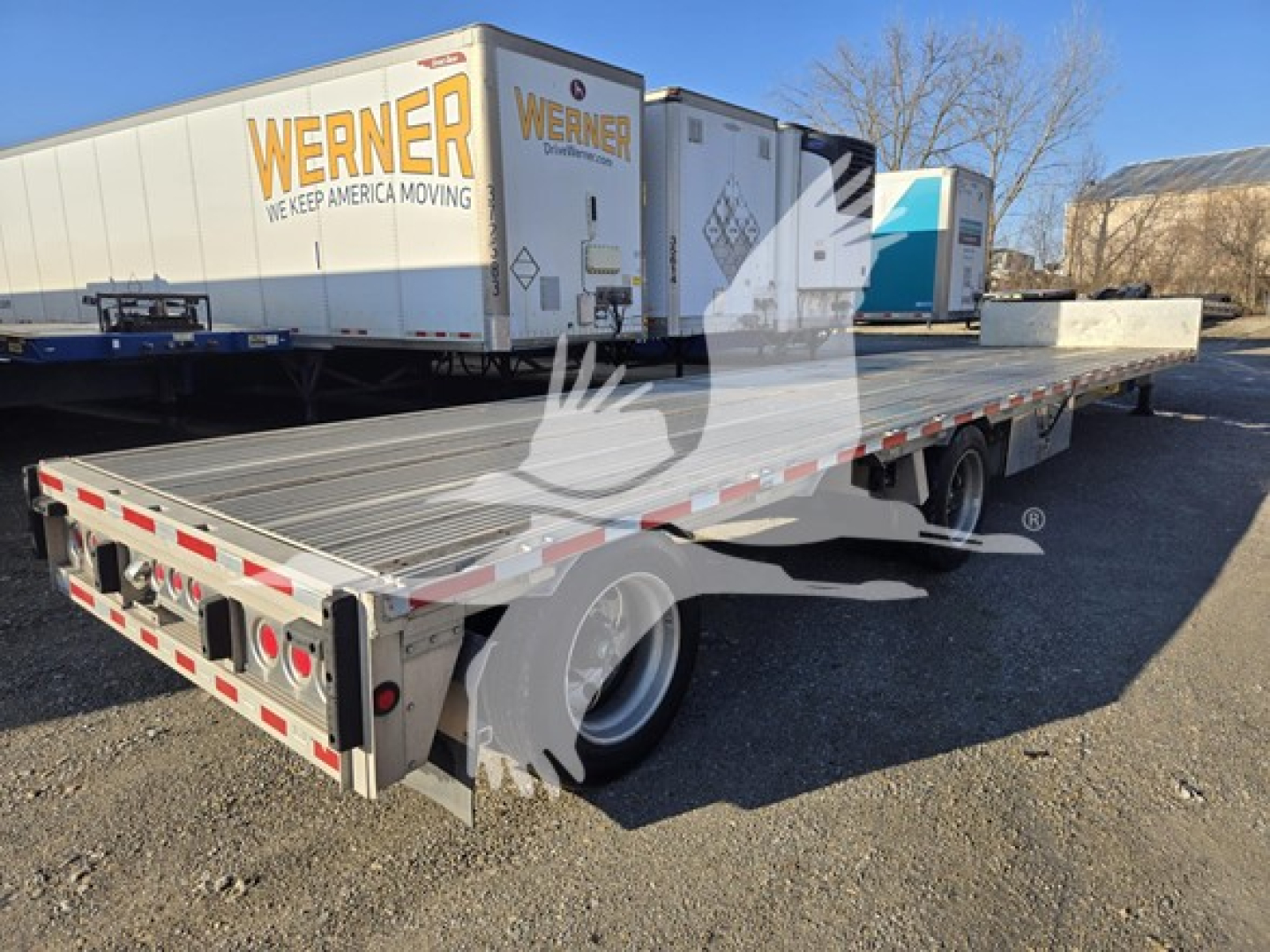 Drop deck trailer with Werner moving logo trailer and reflective edge markings; visible text includes 'WERNER' on the side of a box trailer in the background and 'DROP-DECK' style equipment; no other legible data visible.