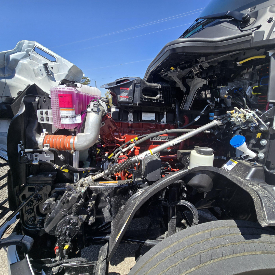 Mack PR64T76M truck engine bay showing coolant reservoir, pink coolant overflow tank, various hoses, wiring, and engine components.
