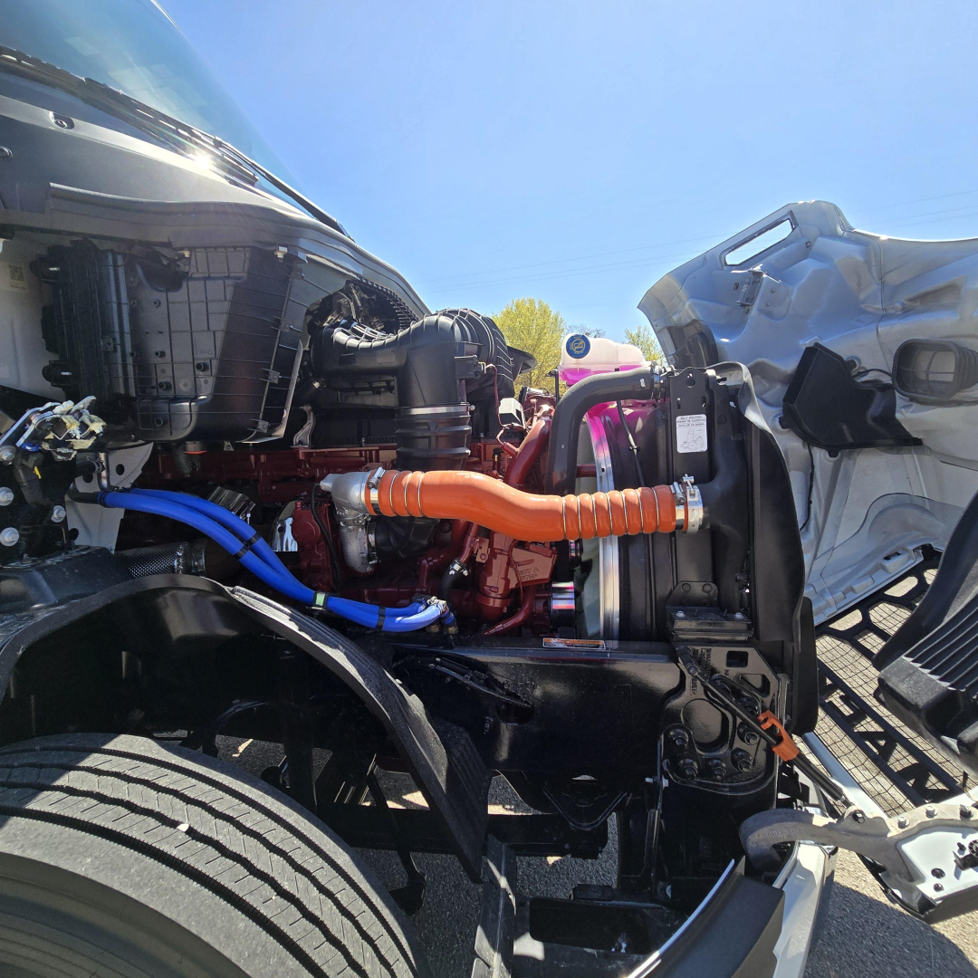 Truck engine bay with orange coolant hose, blue hose, radiator, and air intake visible; Mack PR64T76M engine bay side view