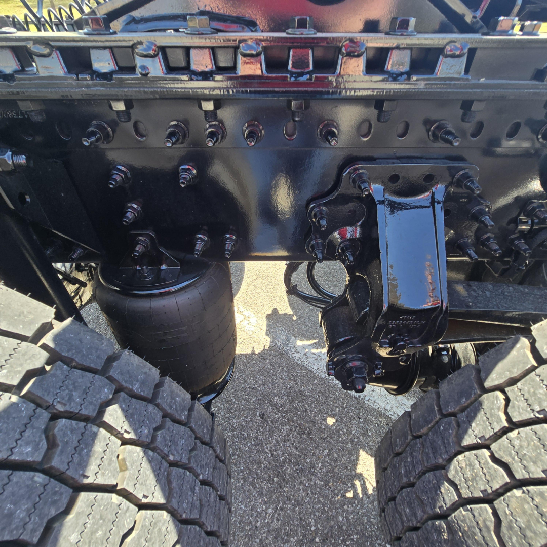 Front axle suspension undercarriage of a Mack PR64T76M truck showing dual rear wheels, air spring (airbag), and leaf spring components with bolts and wiring.