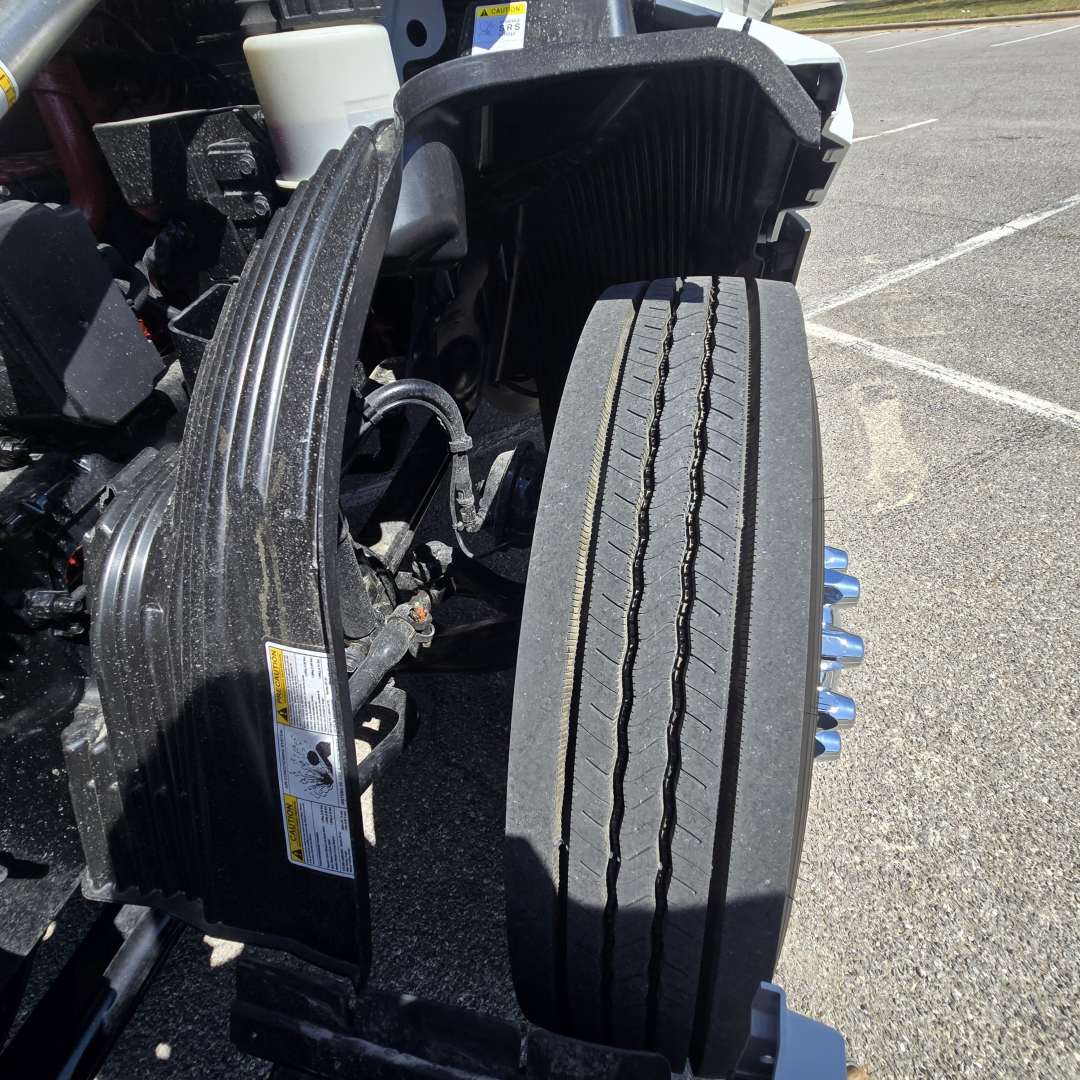 Truck front wheel area of a Mack PR64T76M showing dual tires and engine cooling components with warning labels and a white reservoir in view.