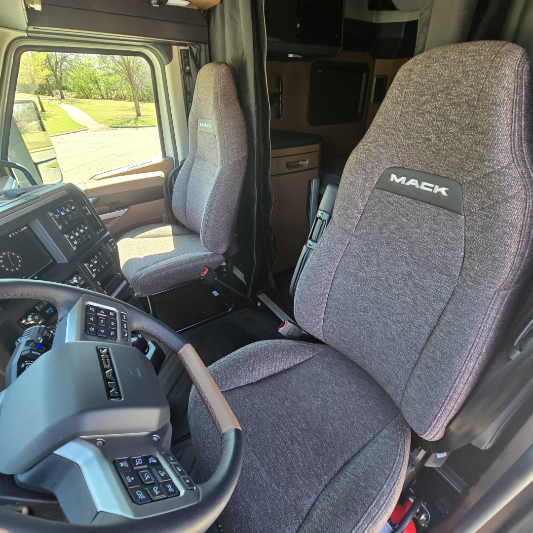 Interior view of a Mack truck cabin showing two fabric seats with ‘MACK’ branding, steering wheel, and dashboard details visible on the left.
