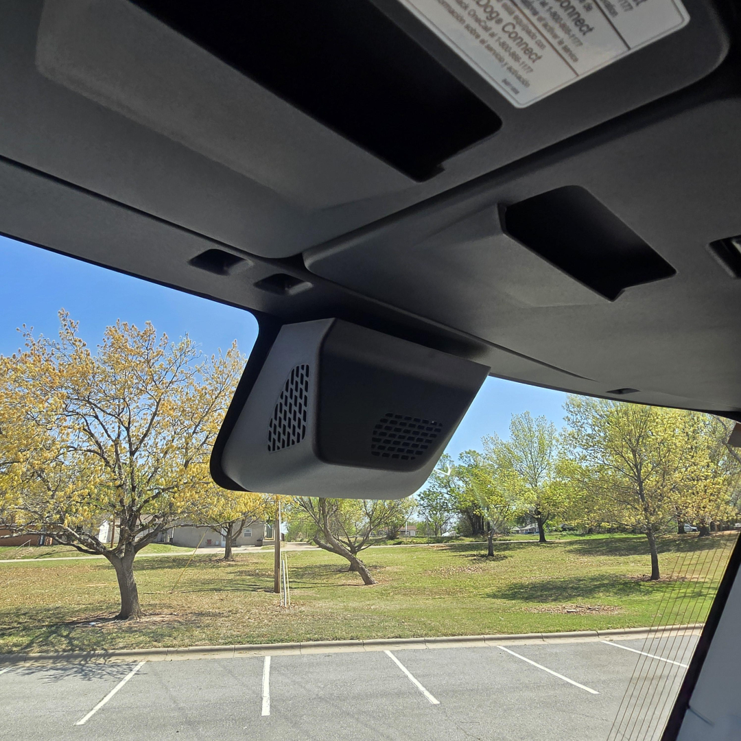 Dashboard view of a 2027 Mack PR64T76M showing overhead display and windshield area with a focus on a mounted interior device (likely sensor/manufacturer hardware) and sunlit exterior lot.