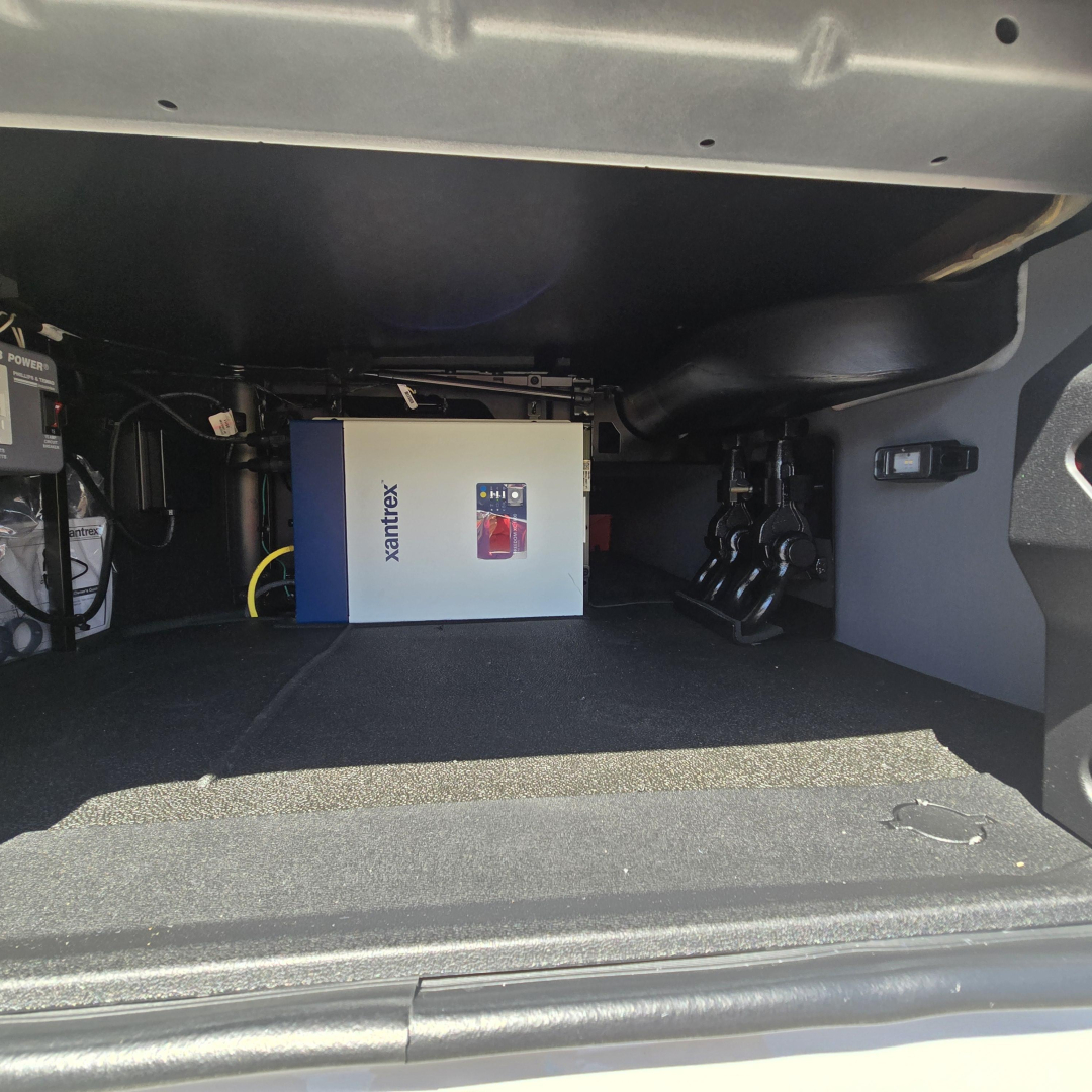 Storage area under a Mack truck sleeper showing an Xantrex inverter/charger box and adjacent cables.