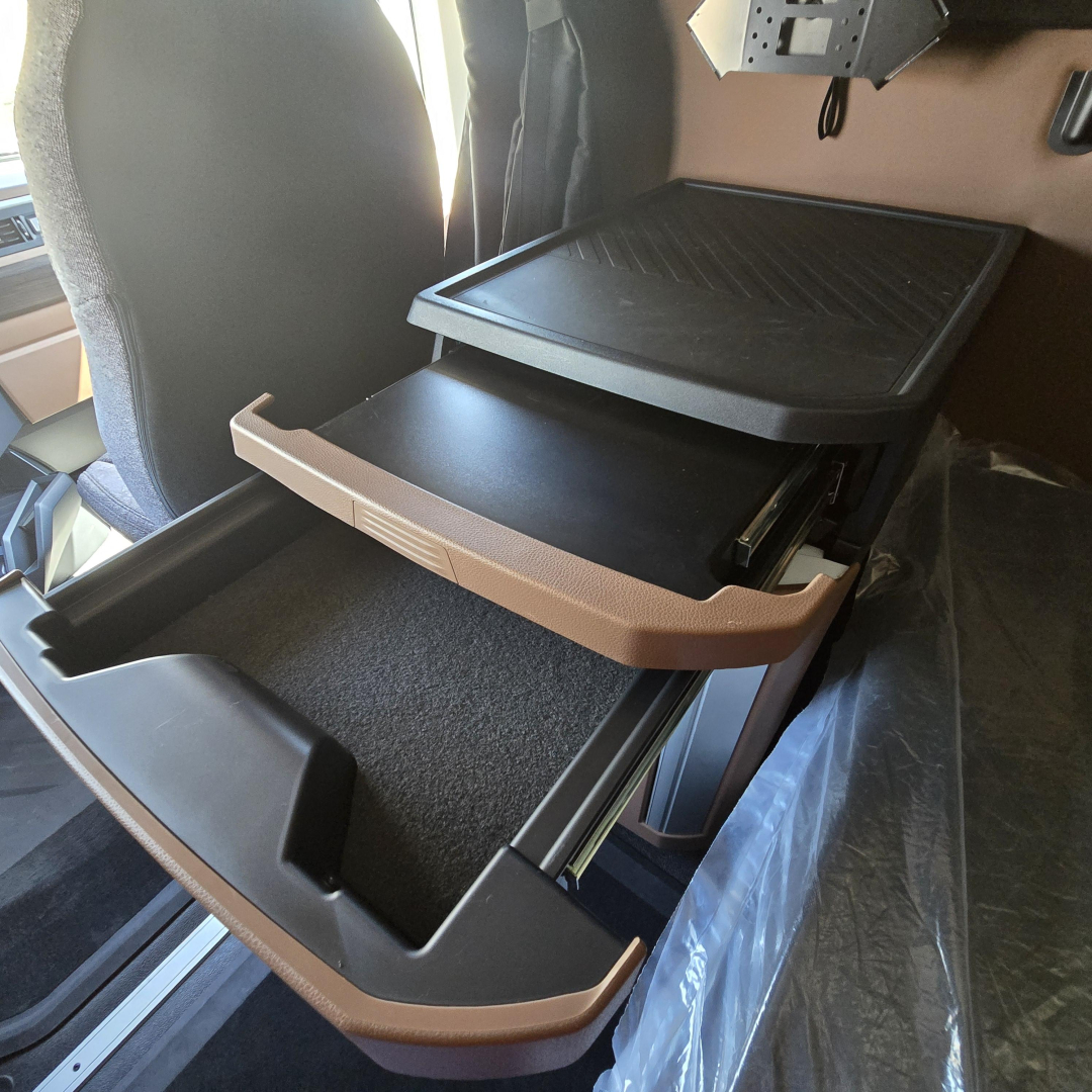 Truck interior showing storage drawer in a Mack PR64T76M sleeper cab, with open tray/compatibility shelf and dash-side console; visibility of cabin seating and control shelf.