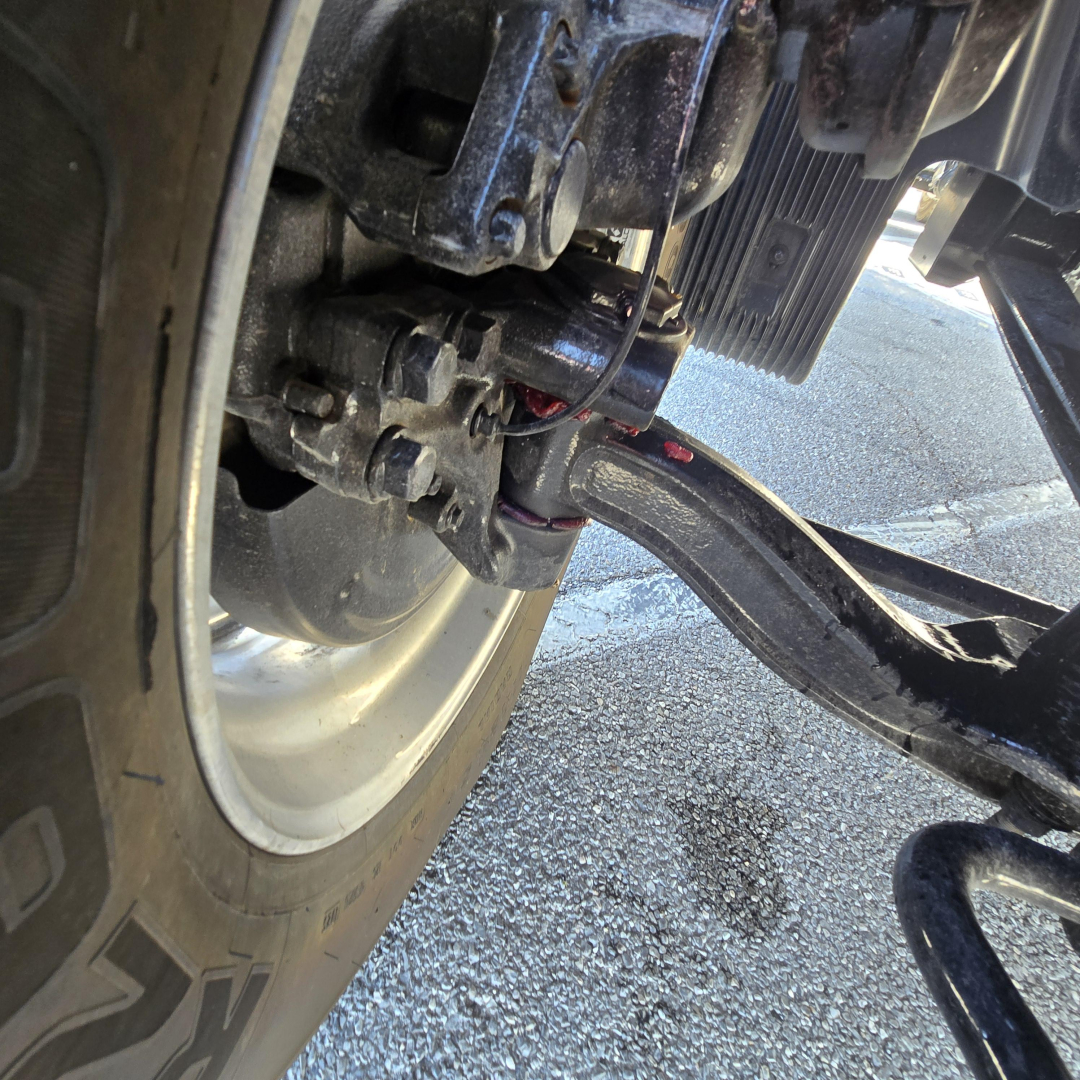Front suspension and drive axle of a new 2027 Mack PR64T76M with visible leaf spring, U-bolts, and leaf spring hanger in a truck yard.