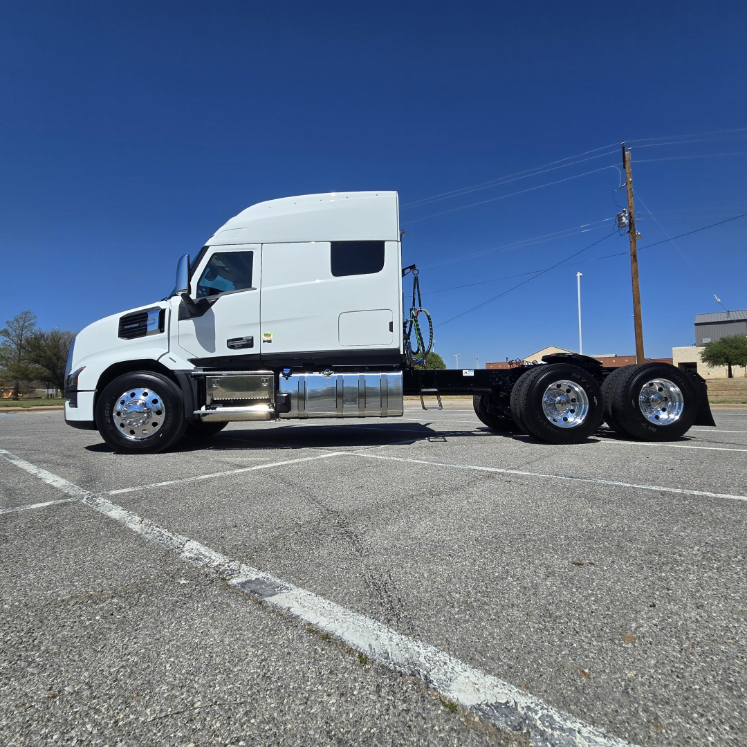 Truck chassis and cab of a white 2027 Mack PR64T76M sleeper with dual rear axles, side fuel tanks, and chrome wheels.