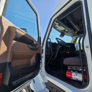 Truck cab interior view with open door shows steering wheel, dashboard, and driver's seat; fire extinguisher mounted under dash; beige door panel; 'MACK' logo on door sill.