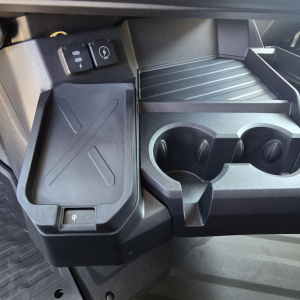 Center console area of a 2027 Mack PR64T76M showing cup holders and a small storage lid with a power/USB panel nearby.