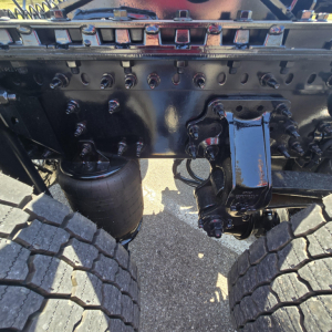 Front axle suspension undercarriage of a Mack PR64T76M truck showing dual rear wheels, air spring (airbag), and leaf spring components with bolts and wiring.