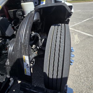 Truck front wheel area of a Mack PR64T76M showing dual tires and engine cooling components with warning labels and a white reservoir in view.