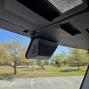 Dashboard view of a 2027 Mack PR64T76M showing overhead display and windshield area with a focus on a mounted interior device (likely sensor/manufacturer hardware) and sunlit exterior lot.