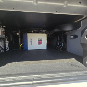 Storage area under a Mack truck sleeper showing an Xantrex inverter/charger box and adjacent cables.