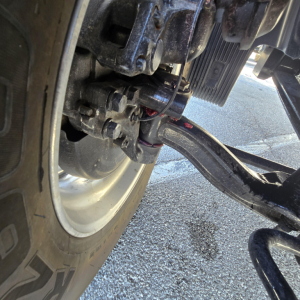 Front suspension and drive axle of a new 2027 Mack PR64T76M with visible leaf spring, U-bolts, and leaf spring hanger in a truck yard.