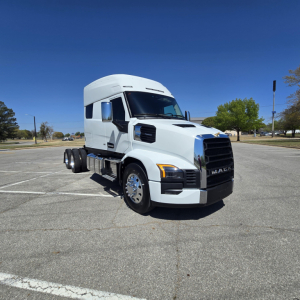 Truck cab in white, front-right view showing Mack PR64T76M with aerodynamic sleeper, dual rear wheels, chrome grille, and daytime parking lot.