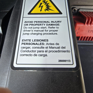 Safety warning label with English and Spanish text on a vehicle component; top shows a yellow triangular hazard symbol and black lightning bolts. Visible text includes: 'AVOID PERSONAL INJURY OR PROPERTY DAMAGE: Do not jump start. Refer to 
