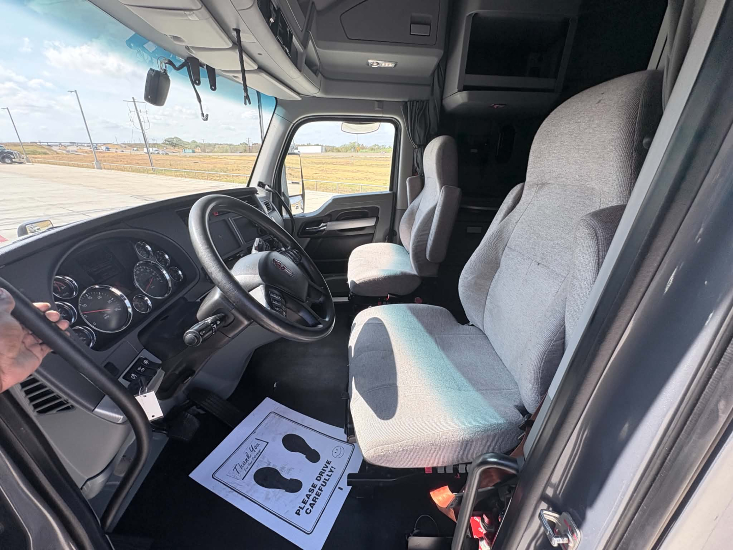Kenworth T680 interior cab showing steering wheel, dashboard, and driver's seat with light gray fabric cushion; view through open door into cabin.
