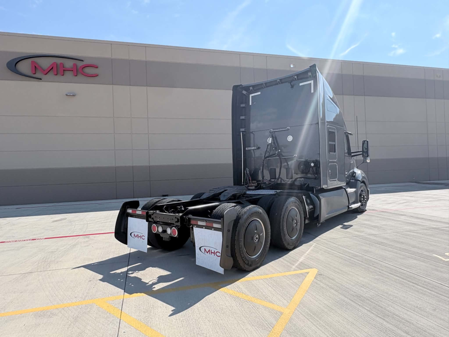 Kenworth T680 tractor in driveway showing rear view with sleeper, dual rear axles, and fifth wheel; MHC logo on building in background.