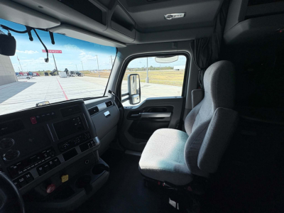 Truck cab interior front view with steering wheel, dashboard, and passenger seat; windshield shows exterior lot and sky.