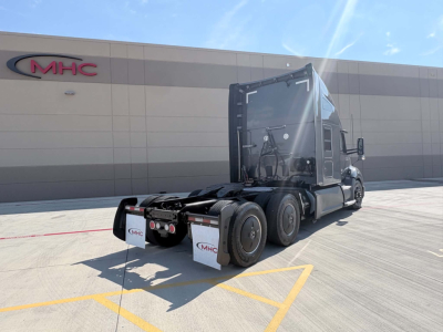 Kenworth T680 tractor in driveway showing rear view with sleeper, dual rear axles, and fifth wheel; MHC logo on building in background.