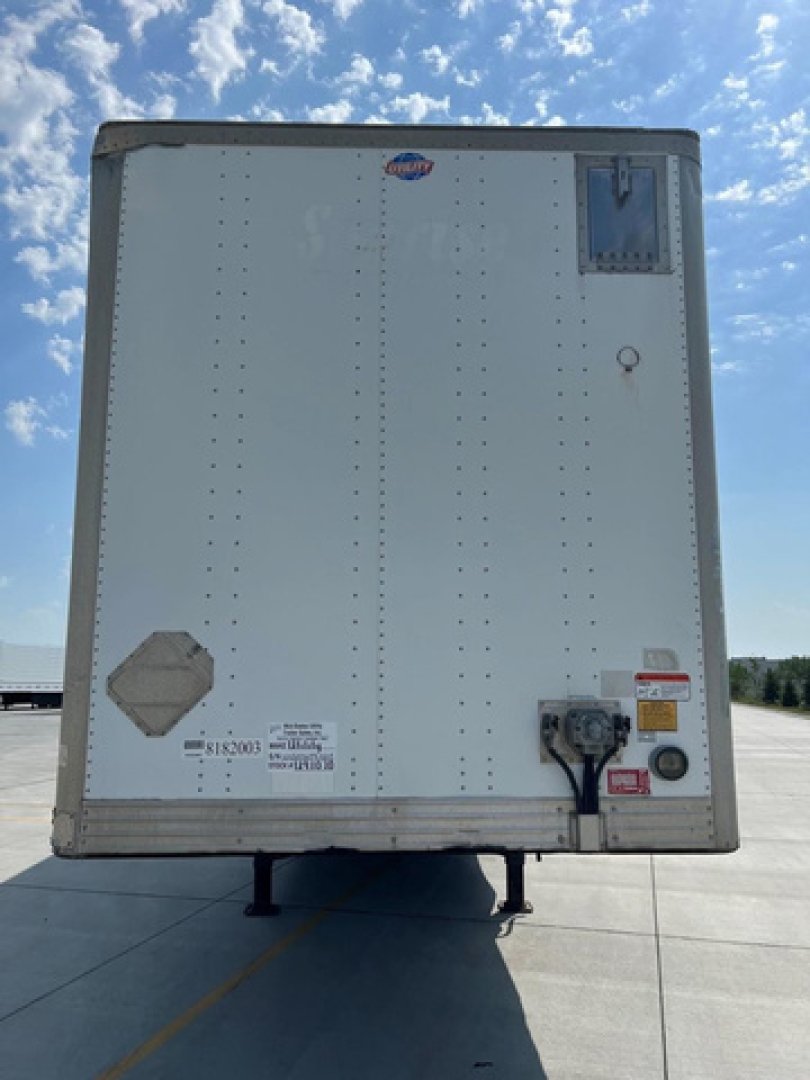 Rear of a white utility trailer showing various decals and electrical outlet with labels. Visible text includes VIN sticker and numbers near bottom left, a small blue and white sticker at top center, and red/black warning stickers near bott
