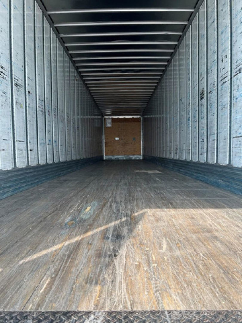 Trailer interior showing empty box trailer with wood deck and metal sidewalls, long rectangular cargo area, rear doorway with wooden end wall.