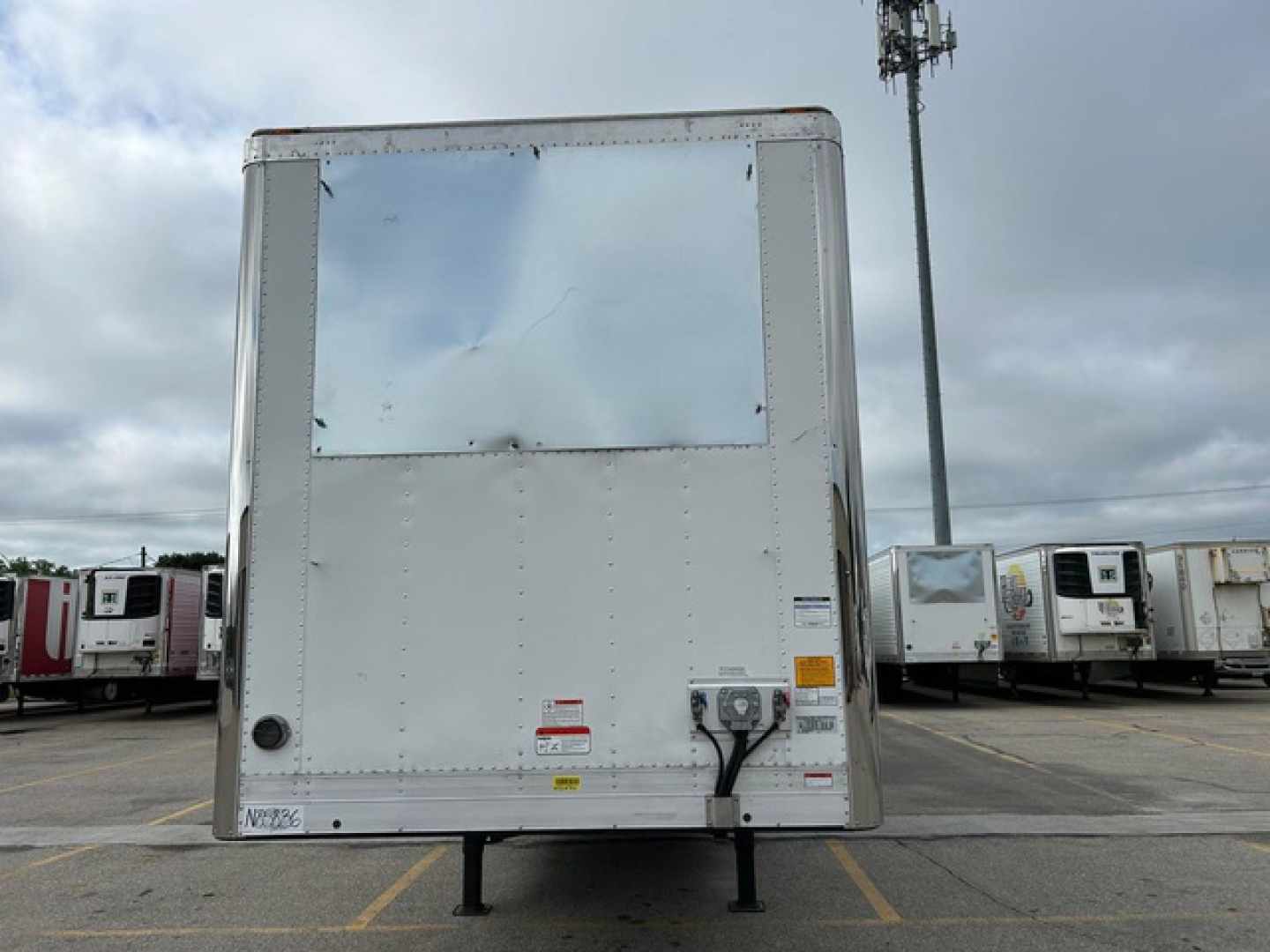 Trailer rear view showing a white refrigerated utility trailer with a reflective window, two entry doors, and support legs. Visible decals and warning labels at lower right.