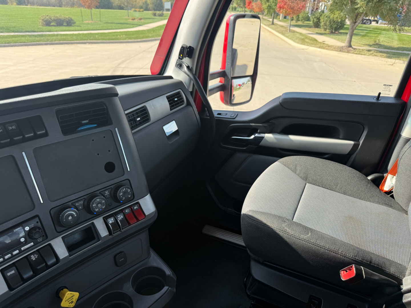 Truck cabin interior of a 2023 Kenworth T680 from the left side, showing the dashboard with assorted controls and a gray cloth passenger seat. Side mirror visible through the window.