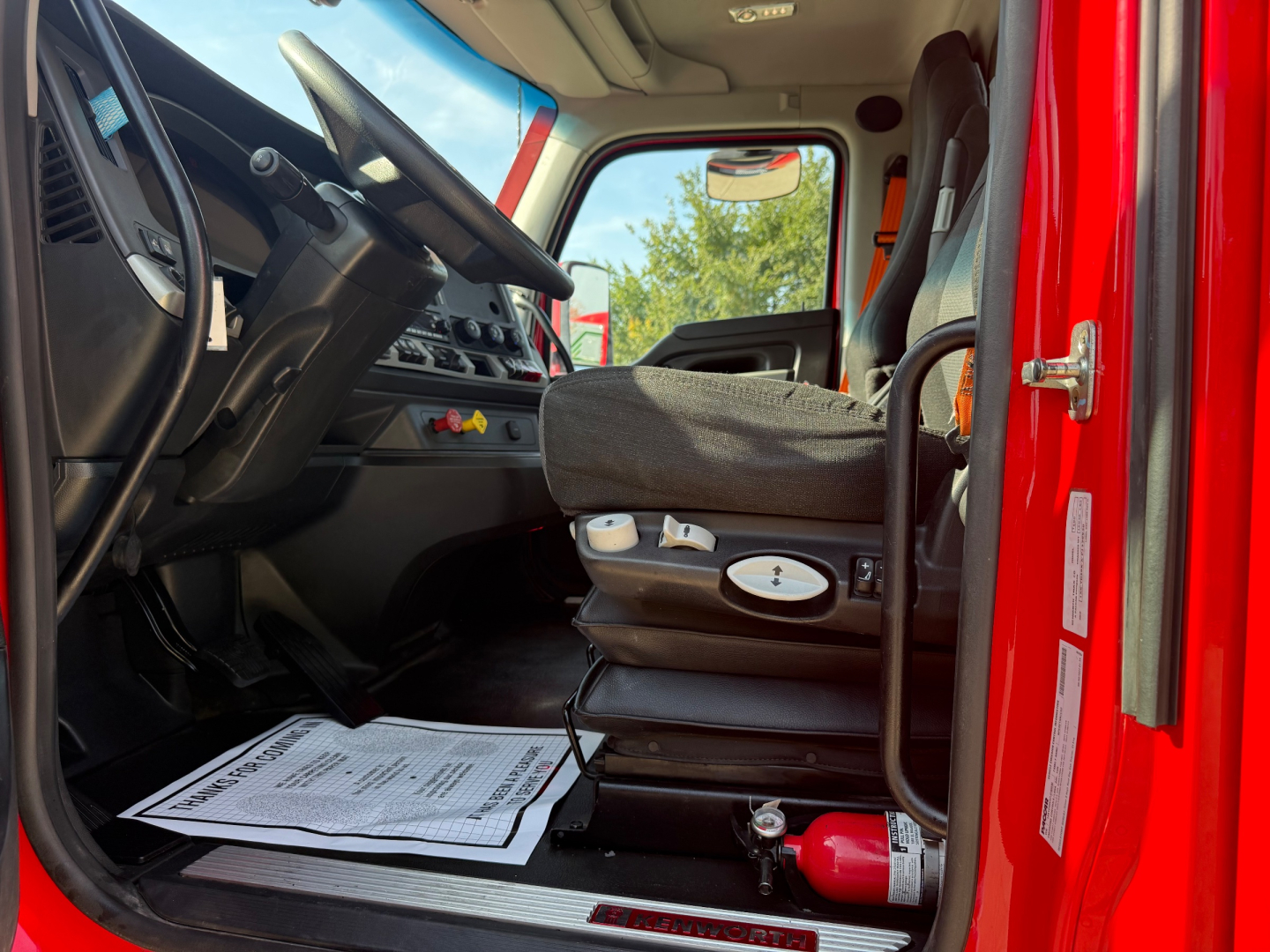 Kenworth T680 truck cabin interior with steering wheel, dashboard, driver seat, floor mat, and a fire extinguisher in the cab; door frame visible.
